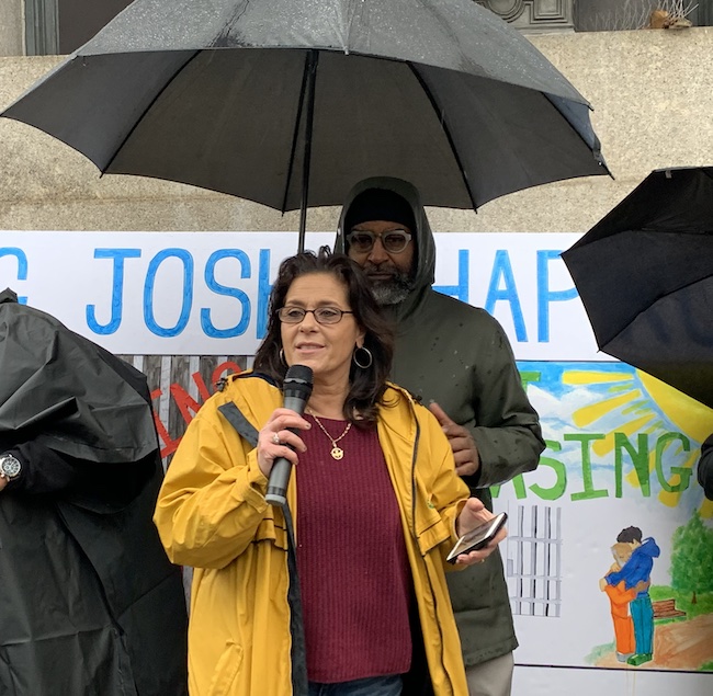 Marcie speaks in a mic while someone holds an umbrella over her head