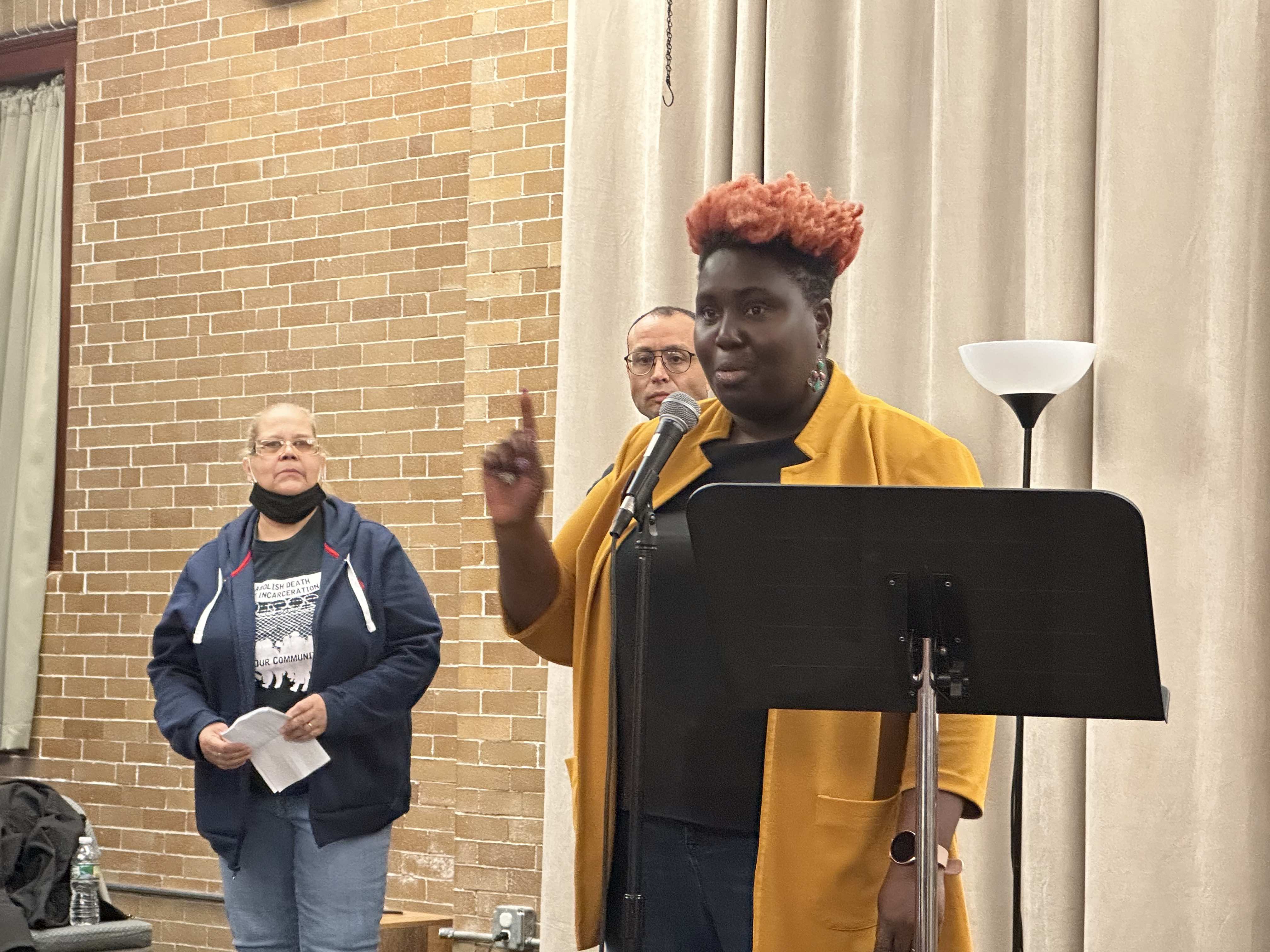 Nikki Grant wears a yellow sweater and holds her finger up while speaking