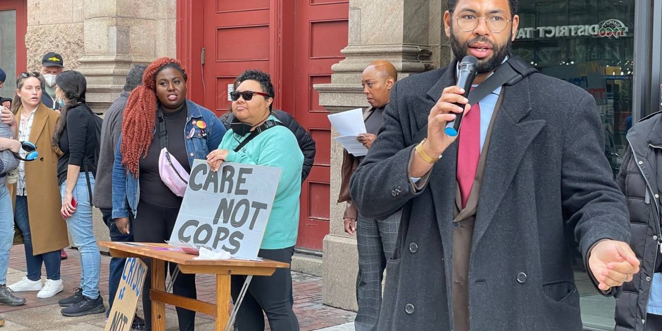 State Rep. Rick Krajewski wears a suit and speaks into the mic as people stand in the background including Kris Henderson holding a 'care not cops' sign