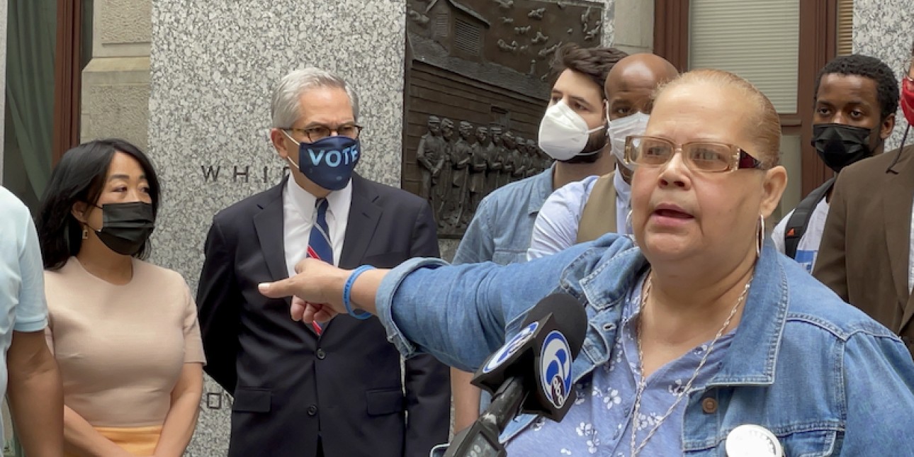 Mrs Dee Dee stands at a podium and points towards Larry Krasner who is standing next to Helen Gym