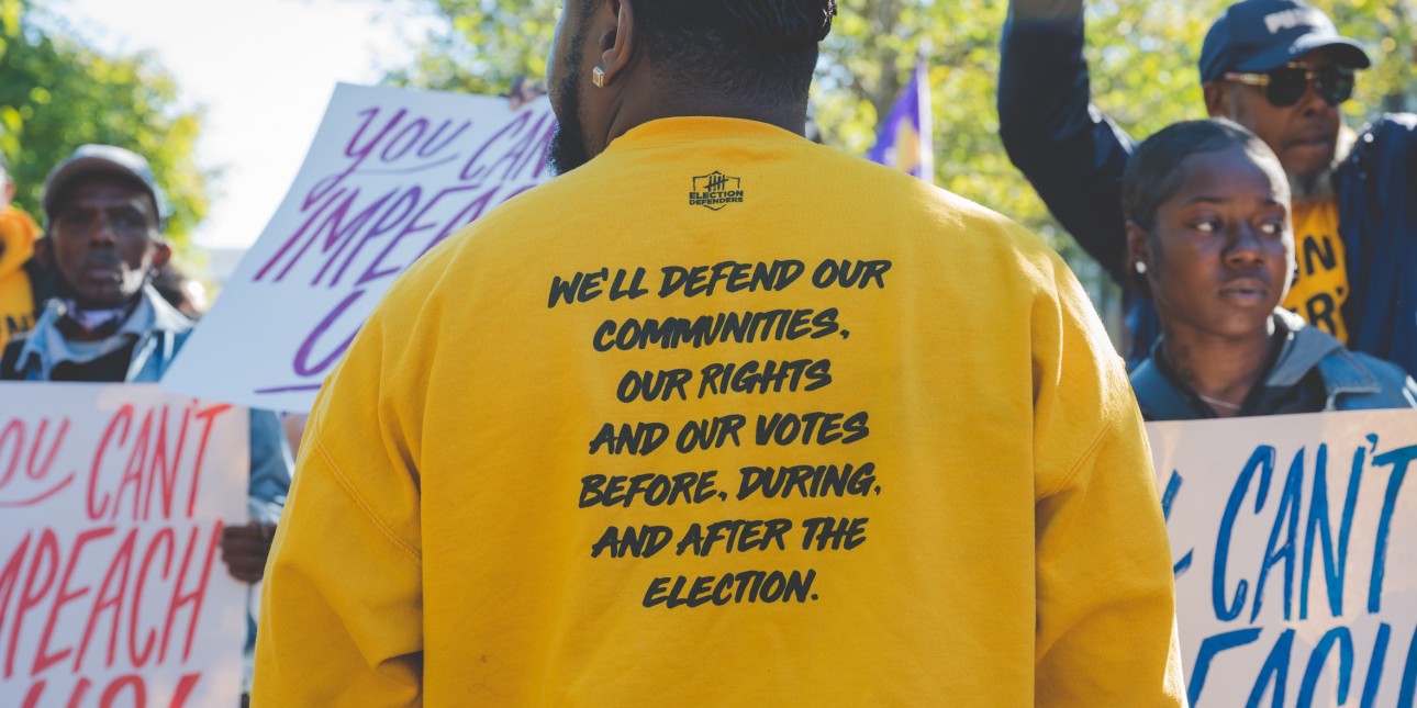 a man wearing a yellow election defenders sweatshirt displays the back of his sweatshirt for the camera