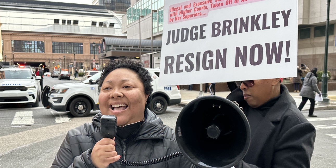 Kris Henderson speaks into a megaphone in front of a sign that reads Judge Brinkley resign now