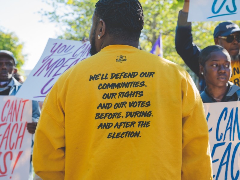 a man wearing a yellow election defenders sweatshirt displays the back of his sweatshirt for the camera