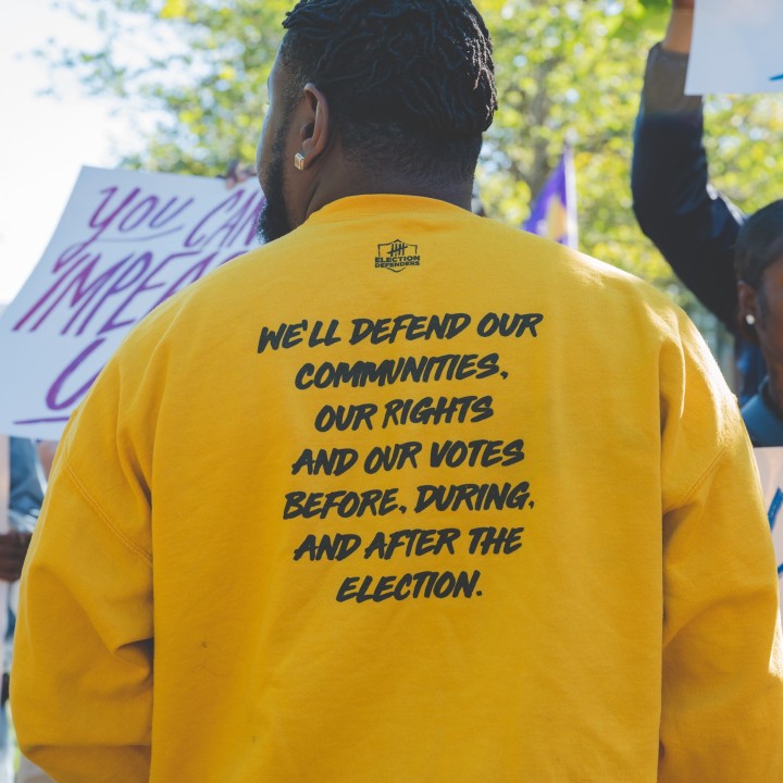 a man wearing a yellow election defenders sweatshirt displays the back of his sweatshirt for the camera