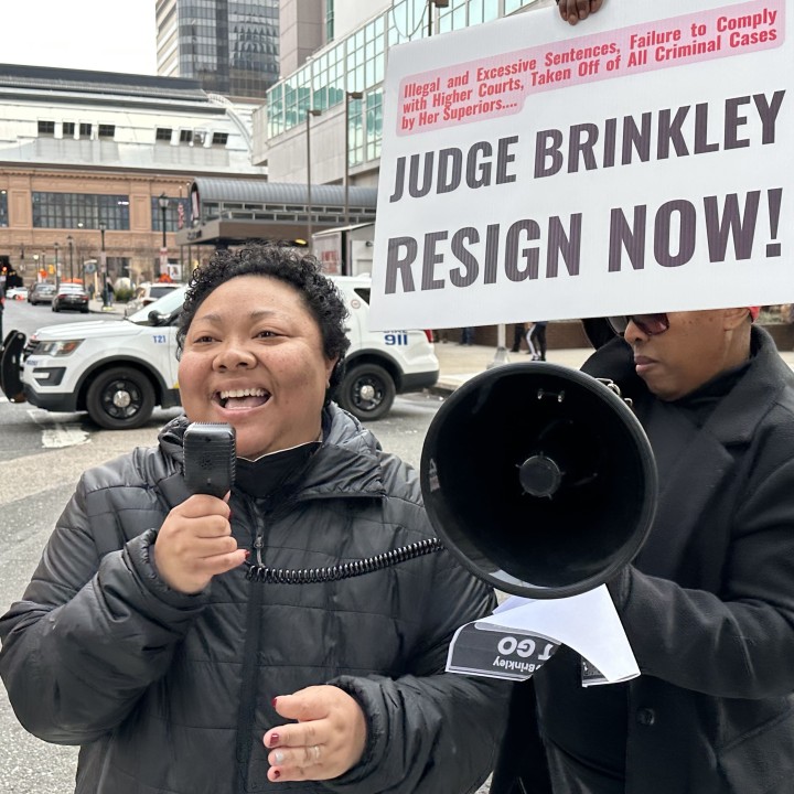 Kris Henderson speaks into a megaphone in front of a sign that reads Judge Brinkley resign now